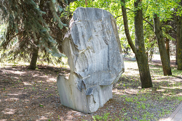 Large granite boulder stands on gravel in a park among trees. The natural object contrasts with the paved path and green foliage.