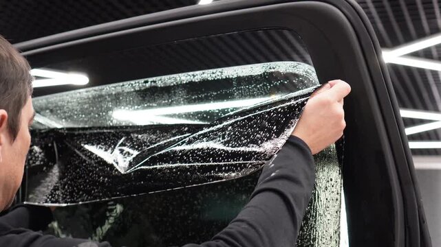 Male car service worker glue window tinting in luxury car detailing car repair the concept of love for cars and protection. Applying tinting foil on a car window in the tuning studio close-up. 