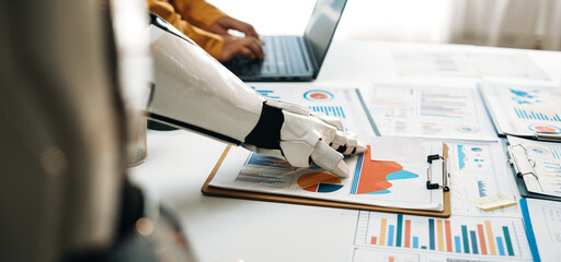 A robot hand interacts with analysis documents on a table while a human works on a laptop, showcasing collaboration in technology and data analysis.Noogenesis