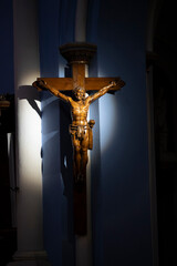 A wooden crucifix stands out against the blue wall of Basilique Notre-Dame-de-Buglose in Saint-Vincent-de-Paul. The light highlights the details of the sculpture, creating a serene atmosphere.