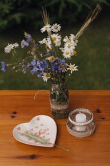 Flowers in a rustic jar alongside a decorative heart plate and candle on a wooden table in a garden setting
