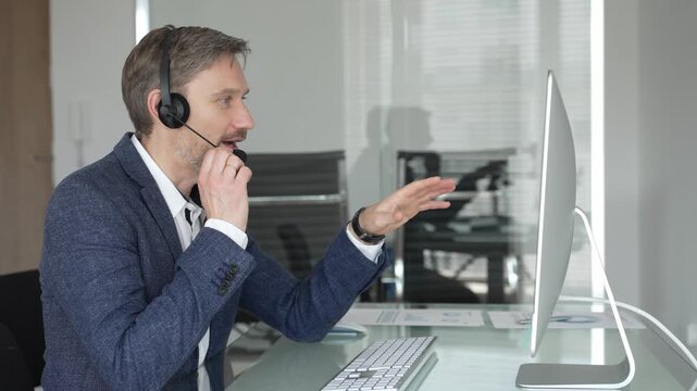 Male call center agent wearing headset providing online customer support, consulting client on headset video call. Business people concept