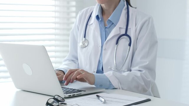 Female doctor physician working at desk, typing on laptop, medical records and pen nearby, bright window with blinds illuminating clinical workspace. Medicine and health care concept