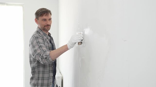 Construction worker wearing gloves and using a putty knife to plaster a white wall, performing home renovation and improvement tasks with precision