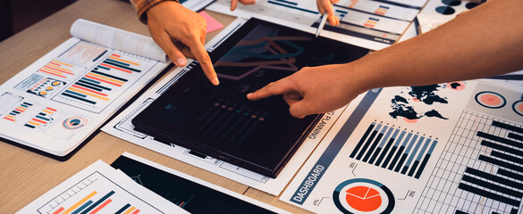 A vibrant business meeting scene depicts multiple hands pointing at data visualizations and charts on reports and a digital tablet, emphasizing teamwork and strategy. SACTR