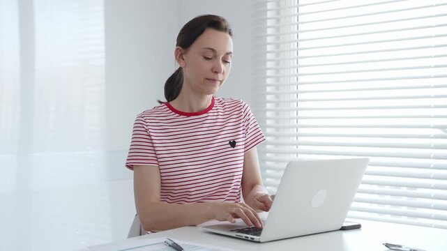 Focused female freelancer working from home, using laptop and sitting at white desk near window with blinds, concentrating on her online work. Business people concept