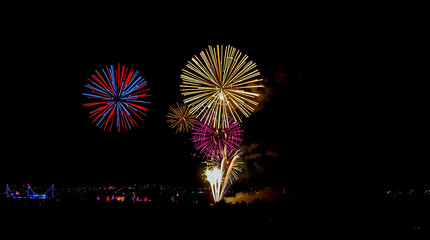 Colorful fireworks explode in the night sky above a distant crowd