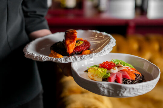 Elegant, gourmet fusion cuisine dishes featuring fish, fresh vegetables, and grains are served by a waiter in a fine dining restaurant setting