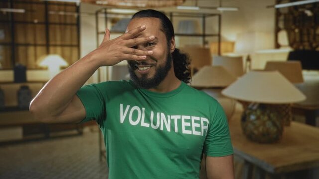 Man covering his eyes with his hand while smiling in a lamp shop building, wearing a green volunteer shirt; playful charity.