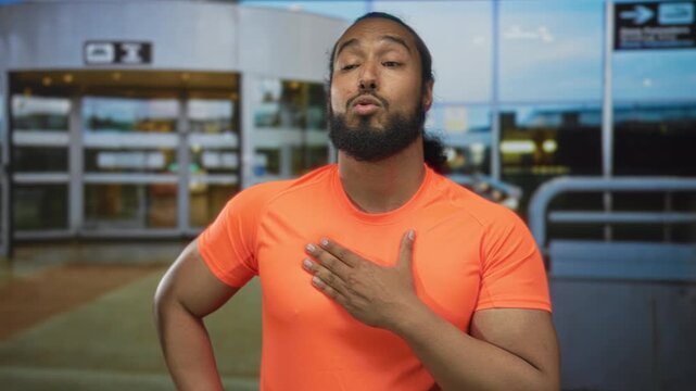 Man with hand on chest exhaling at airport terminal outdoors in bright orange tshirt, eyes closed and one hand on hip; relief recovery.