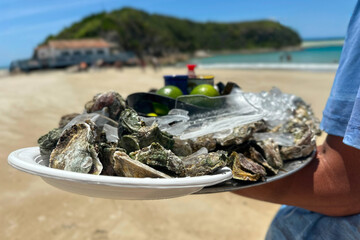 Close-up view of fresh oysters served on a tray with crushed ice and whole limes, held by a vendor on a sunny beach.