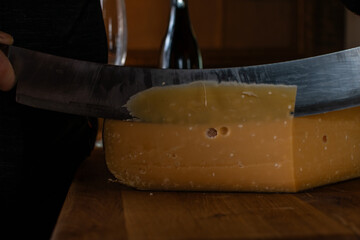 A person is slicing a thick piece of aged hard cheese with a sharp kitchen knife on a wooden cutting board. Close-up on the moment of cutting. Food preparation concept