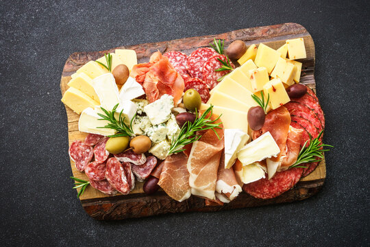 Italian appetizers. Cheese and meat - jamon, salami with olives at wooden serving board at black background. Top view.
