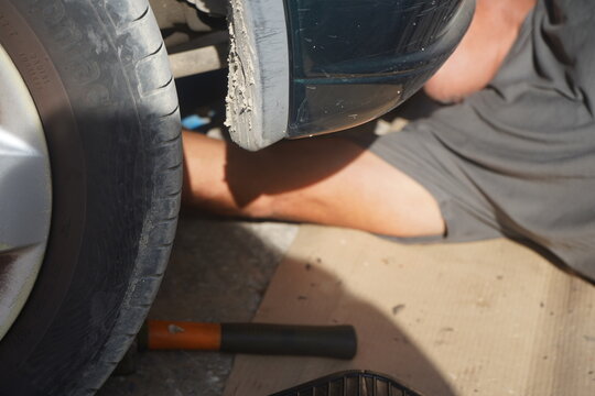 A person wearing a grey shirt is lying on the ground, working on the undercarriage of a car, seen next to the tire and mudguard. Focus on car repair and outdoor maintenance.