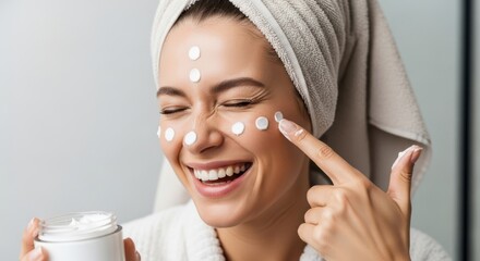 A happy woman with a towel on her head applies cream to her face, smiling with her eyes closed, holding a jar of moisturizer
