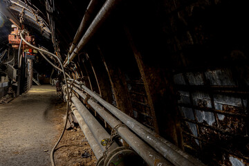 Heavy tools and equipment inside an underground mine, with steel chains, pneumatic hoses, and supply lines in active use. The scene shows industrial work conditions, mining technology, and harsh subte