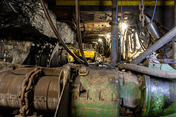 Heavy tools and equipment inside an underground mine, with steel chains, pneumatic hoses, and supply lines in active use. The scene shows industrial work conditions, mining technology, and harsh subte