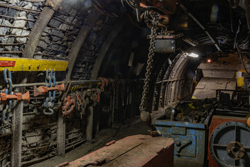 Heavy tools and equipment inside an underground mine, with steel chains, pneumatic hoses, and supply lines in active use. The scene shows industrial work conditions, mining technology, and harsh subte