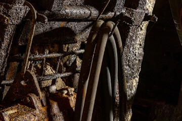 Heavy tools and equipment inside an underground mine, with steel chains, pneumatic hoses, and supply lines in active use. The scene shows industrial work conditions, mining technology, and harsh subte