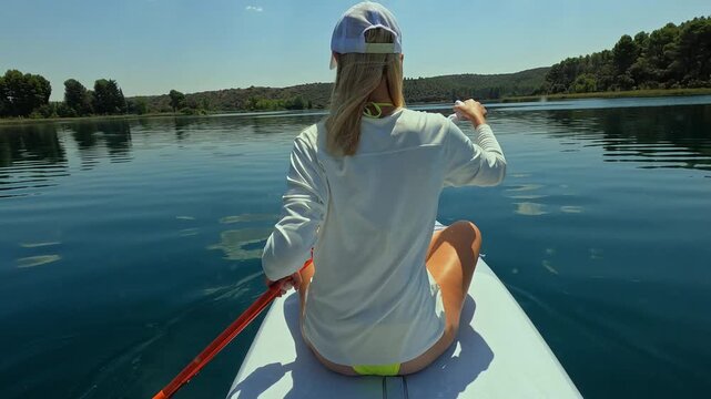 Young athletic woman with long blonde hair paddling on paddleboard on beautiful calm lake during sunny summer day, enjoy serene nature and favorite water sport activity