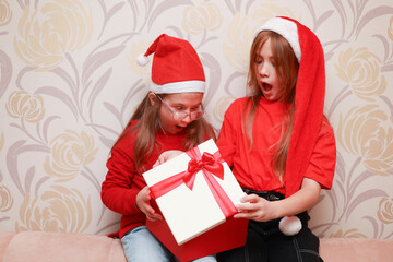 Girls in New Year's looks with a gift are sitting at home on the sofa.