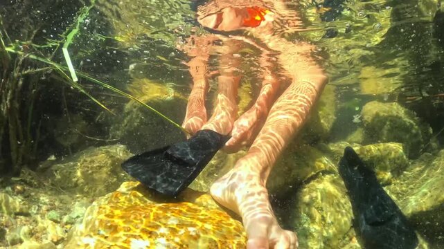 Underwater view of young person sitting on rocks in clear water and putting on black fins, getting ready for snorkeling adventure with sun rays creating caustic patterns on skin