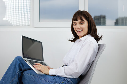 A woman sits in a bright modern office with a laptop on her lap, smiling at the camera. The scene conveys a calm, productive workspace and casual professional vibe.