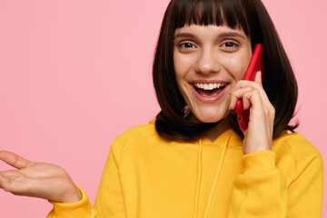 A cheerful young woman in a bright yellow hoodie chats on a red phone, smiling against a pink backdrop. Casual portrait conveying friendly communication and positive energy.