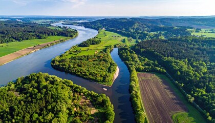 Aerial View Of A Wide River Meandering Through Lush Green Forests And Agricultural Fields Under A Bright Blue Sky