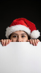 A child in a Santa hat peeks over a white surface with wide, curious eyes. Small hands grip the edge as the bright hat contrasts with the dark background in a festive, playful scene.