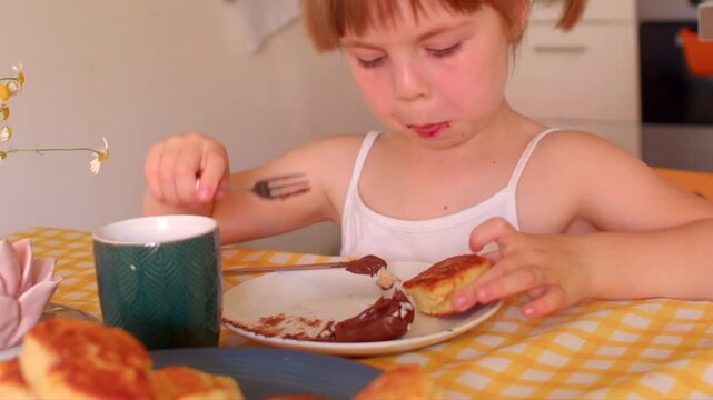 toddler tasting pancake at kitchen table, sticky chocolate syrup on chin, yellow gingham tablecloth, green mug