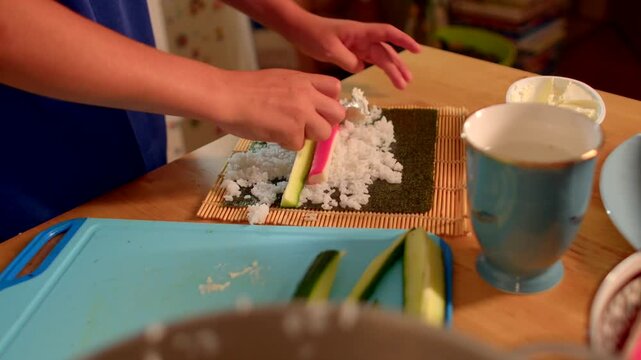 white child placing cucumber and crab stick onto sushi rice on nori sheet, careful small hands arranging