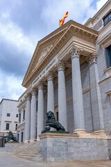 The neoclassical facade of the Spanish Congress of Deputies features six columns, a pediment, steps, and two large bronze lion statues flanking the main golden doorway