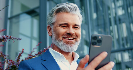 Adult businessman stands in front of modern glass office building, holding smartphone in front of him while having an online video call with colleague outdoors.