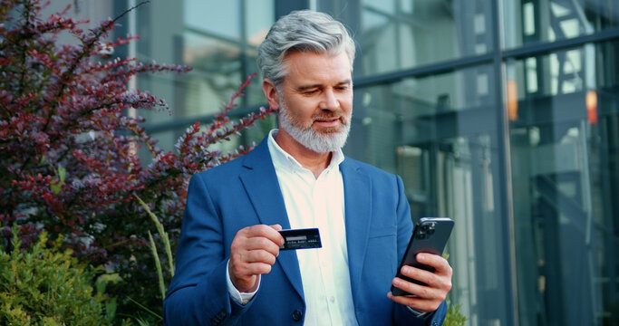 Adult man using smartphone and credit card for online banking while sitting near a glass office building. Concept of fintech, digital transactions, and mobile money management