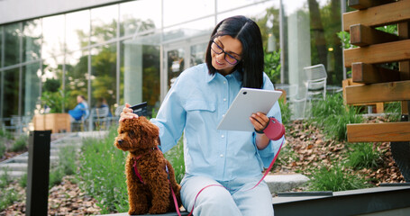 Young woman in glasses wearing casual clothes, with an earbud in ear, entering credit card information for an online purchase on tablet while pet dog sits calmly on leash nearby in green park