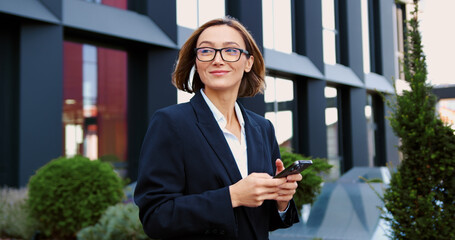 Business woman employee chatting online, texting using a smartphone walking on the street near an office building corporate office typing text messages on mobile phone checking emails successful