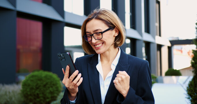 Businesswoman employer in suit celebrate euphoric news amazing receive message on mobile phone good news. Woman reading good news on smartphone standing on city street. Concept of success and