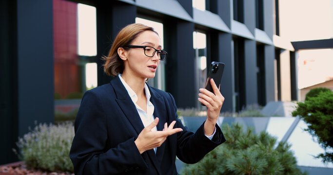 Business woman in glasses walking and having a video call on smartphone in urban business district waving hand. Female entrepreneur having an online meeting with client or business partner.