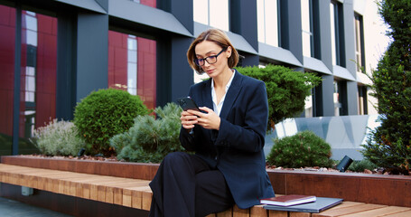Business woman using smartphone outdoors standing on city street, looking to mobile phone screen, enjoying urban atmosphere. Cheerful female using mobile application, texting message, shopping online.