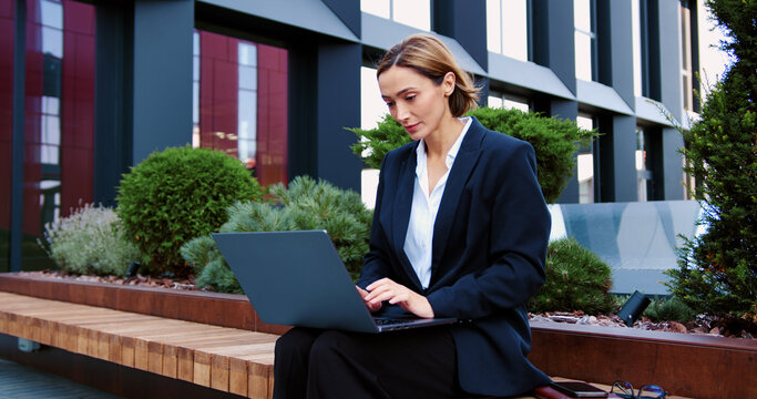 Smiling woman in suit using laptop in modern outdoor office environment. Female freelancer typing at computer keyboard chatting online with a partner or a client outdoors. Confident blonde focused on