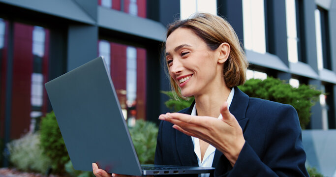 Portrait of smiling woman using digital laptop looking at screen of notebook speaking during online video call on social media while sitting on bench on city street.