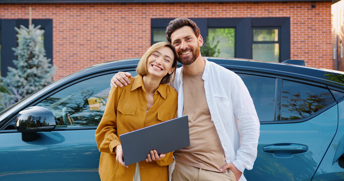 Smiling businessman and businesswoman with laptop standing near electric car outdoors, looking at camera and posing together.