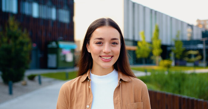 Beautiful young woman with natural smile, dressed in business casual attire, walks through sunny city park during her lunch break, looking directly at the camera with friendly and confident expression