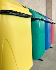 Row of colorful recycling bins used for waste separation in a public area. The image shows yellow, green, blue, and red containers lined up against a concrete wall.