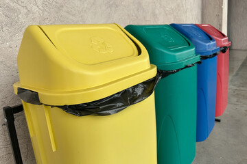 Row of colorful recycling bins used for waste separation in a public area. The image shows yellow, green, blue, and red containers lined up against a concrete wall.