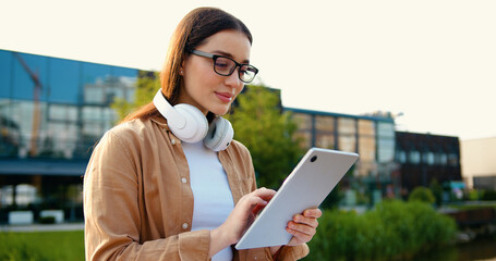 Portrait of young woman in city park with headphones around neck and glasses, working on tablet on commercial building background, outdoors