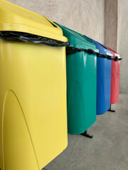 Row of colorful recycling bins used for waste separation in a public area. The image shows yellow, green, blue, and red containers lined up against a concrete wall.