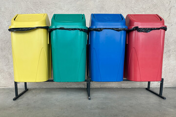 Row of colorful recycling bins used for waste separation in a public area. The image shows yellow, green, blue, and red containers lined up against a concrete wall.