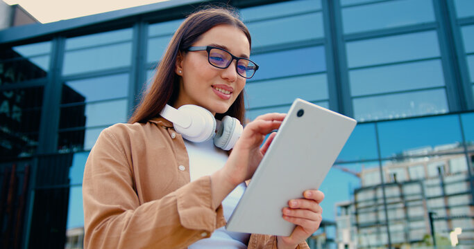 Close-up of smiling young woman wearing headphones around her neck, using tablet while standing in front of a modern office building, outdoors